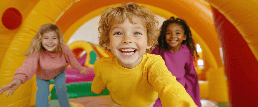 Trois enfants heureux rient et jouent joyeusement dans un château gonflable multicolore. Un garçon blond au premier plan sourit à la caméra.