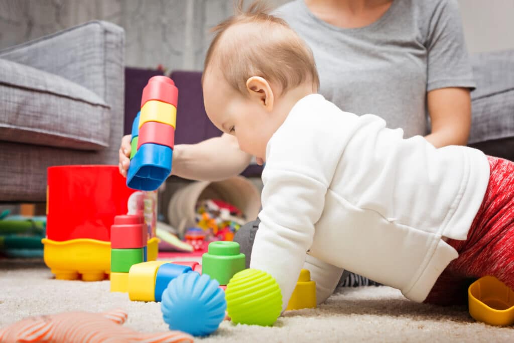 Bébé en body blanc et pantalon rouge, rampant sur un tapis avec des blocs colorés. Un adulte joue avec lui, créant une tour.
