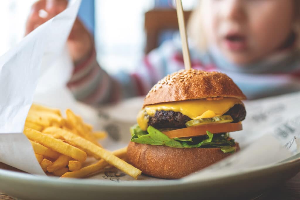 Gros plan sur un cheeseburger avec frites sur une assiette. Un enfant blond, la bouche ouverte, regarde le repas avec curiosité dans un restaurant.