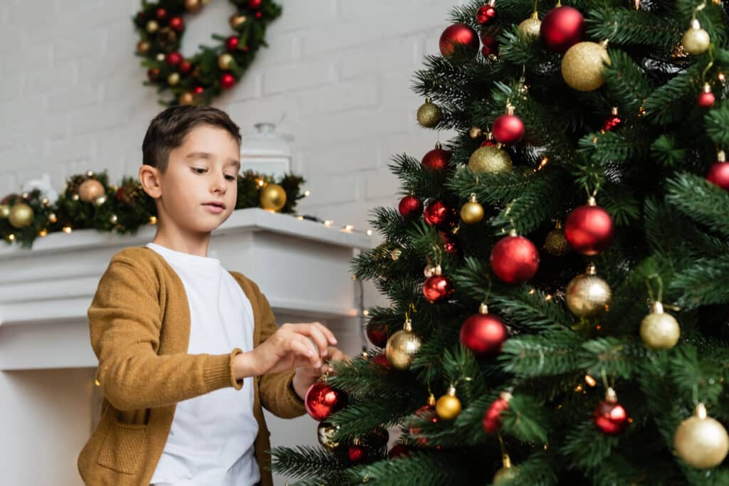 Jeune garçon décore le sapin de Noël avec un ornement rouge, entouré de lumières et décorations festives.