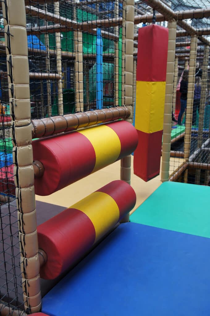 Parcours d'aventure pour enfants avec rouleaux à escalader rouges et jaunes, poteaux matelassés beiges et filets de sécurité.